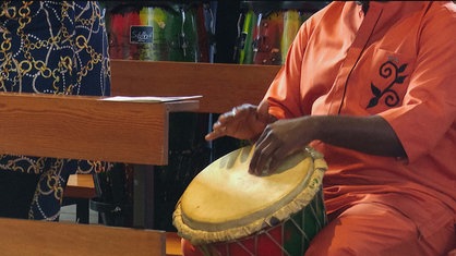 Ein Mann in einem orangen Gewand schlägt mit seinen Händen auf eine afrikanische Trommel  