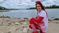 Frau in rot-weißem Karnevalskostüm hockt am Strand von Köln-Rodenkirchen