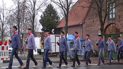 Männer in blau-weißen Hemden laufen mit Holzschuh und Stock mit Wurst über die Straße (Archiv)