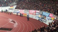 Zu sehen ist eine Tribüne in einem Stadion. An der Tribüne sind viele Fahnen befestigt, mittendrin auch die Ratingen-Fahne. 