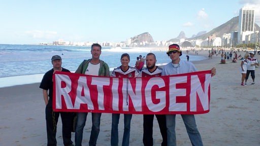Zu sehen sind fünf Männer, die eine Ratingen-Fahne in der Hand halten. Sie stehen dabei an einem Strand.
