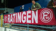 Ein Mann beugt sich in einem Stadion über eine Brüstung. Über der Brüstung hängt eine rote Fahne mit dem Schriftzug Ratingen.
