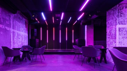 Zu sehen ist ein Raum, der in einem violetten Licht. Es stehen ein paar Tische und Sessel im Raum und es gibt eine kleine Bühne.