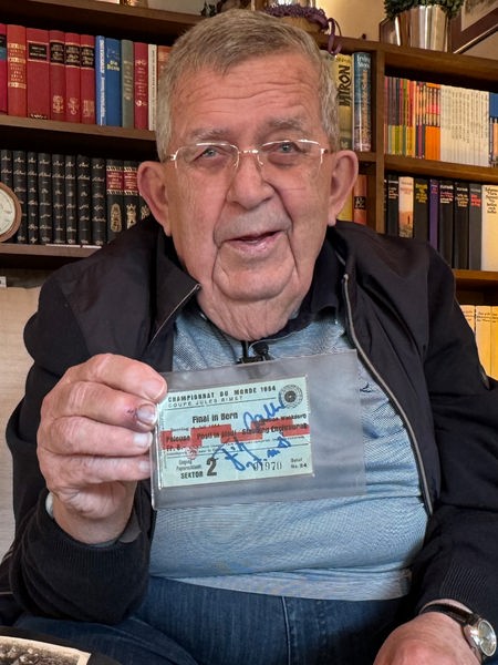 Ein älterer Mann sitzt auf einem Sofa und hält eine alte Eintrittskarte vor sich und lächelt in die Kamera