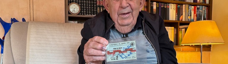 Ein älterer Mann sitzt auf einem Sofa und hält eine alte Eintrittskarte vor sich und lächelt in die Kamera