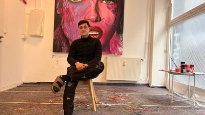 Junger Mann sitzt auf Hocker, im Hintergrund großes Kunstwerk an der Wand