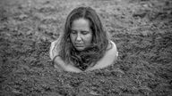 Schwarzweiß Aufnahme: Eine Frau ist bis zu ihren Schultern in Sand eingegraben, die Arme schling sie um ihren Oberkörper