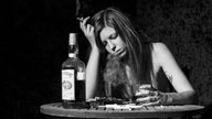 Schwarzweiß Aufnahme: Frau mit langen Haaren sitzt an einem Tisch, stützt Kopf auf eine Hand ab, in der sie eine Zigarette hält, in der anderen Hand hält sie ein Glas mit einer alkoholischen Flüssigkeit darin