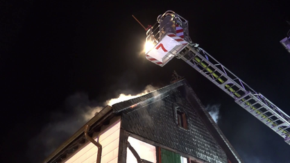 Ein Dachstuhl, aus dem Rauch kommt. Darüber schwebt eine Drehleiter, auf dem mehrere Feuerwehrleute stehen.