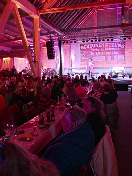 Volle Veranstaltungsaal, Menschen sitzen an langen Tischen. Auf der Bühne steht Ingrid Kühne und erzählt Witze.
