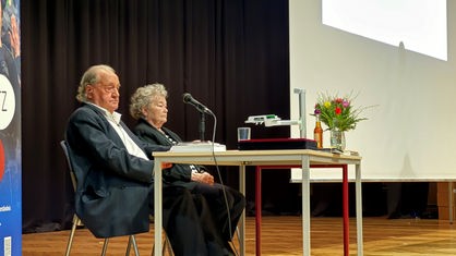 Ivar Buterfas-Frankenthal sitzt an einem Tisch, vor ihm ein Mikro. Rechts neben ihm sitzt seine Frau. 