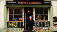 Mann mit Wollmütze auf dem Kopf steht vor einem kleinen Club, über dem Eingang steht der Name "HOTEL EUROPA"