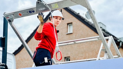 Eine junge Frau in rotem Pulli und weißem Sicherheitshelm auf dem Kopf steht konzentriert auf einer Hebebühne