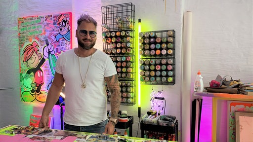 Ein Mann mit Brille und rosa Haar steht in einem Künstleratelier, die Wand ist bunt beleuchtet.