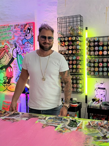 Ein Mann mit Brille und rosa Haar steht in einem Künstleratelier, die Wand ist bunt beleuchtet.