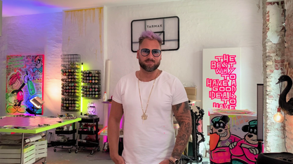 Ein Mann mit Brille und rosa Haar steht in einem Künstleratelier, die Wand ist bunt beleuchtet.