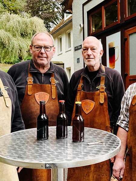 Vier Männer in Lederschürzen stehen an einem Stehtisch. Auf diesem stehen drei Flaschen.