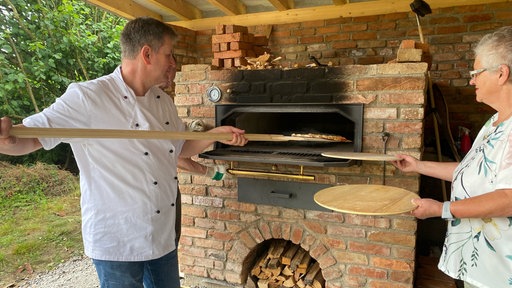 Ein Mann in weißer Kochjacke und eine Frau mit kurzem grauen Haar stehen vor einem großen Backofen aus Backsteinen