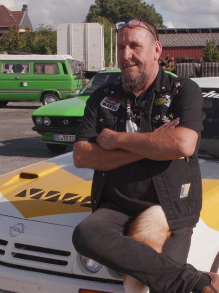"Manta-Jörg" sitzt auf seinem Auto, das weiß lackiert ist mit gelb-grauen Streifen