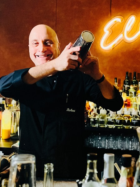 Ein mittelalter Mann mit Glatze, der einen silbernen Cocktailshaker in der Hand hält. Im Hintergrund eine Bar und ein gelber Ellington-Neonschriftzug 