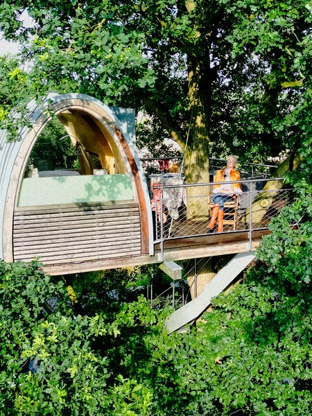 Ein Baumhaus in einer Baumkrone. Auf der Terrasse sitzt eine Frau und sonnt sich.