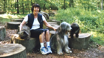 Frau sitzt auf einem Baumstumpf in einem Wald, drei Hunde stehen um sie herum 