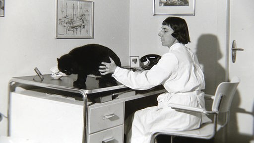 Schwarz-Weiß-Foto: Frau mit dunklem Bob und weißem Kittel berührt schwarze Katze auf Unteruschungstisch