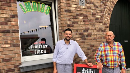 Zwei Männer, die vor einem Imbiss stehen. Der Mann links trägt eine Brille und ist auf eine Tafel gestützt.