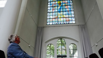 Ein Pfarrer sitzt auf einer Kirchenbank vor einem bunten Kirchenfenster in der Kreuzeskirche in Essen