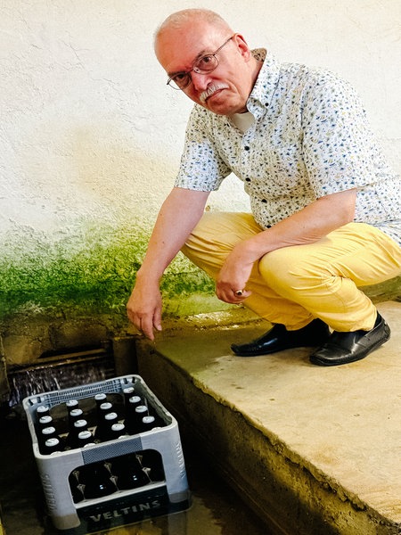 Ein Mann in gelber Hose und gemustertem Hemd hockt vor einem Bierkasten, der im Wasser steht