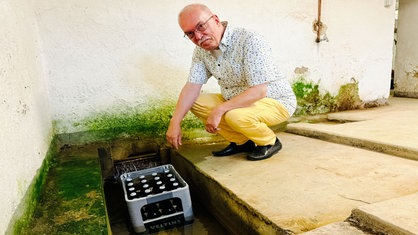 Ein Mann in gelber Hose und gemustertem Hemd hockt vor einem Bierkasten, der im Wasser steht