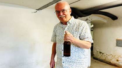 Ein älterer Mann mit Brille und weißem Schnurrbart hält lächelnd eine Bierflasche