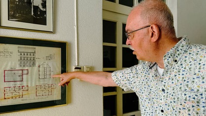 Ein Mann in gemustertem Hemd zeigt auf die Zeichnung eines Haus-Grundrisses