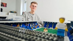 Ein junger Mann in grauem Pulli und kurzen blonden Haaren sitzt hinter einem aus Lego gebauten Fußballstadion