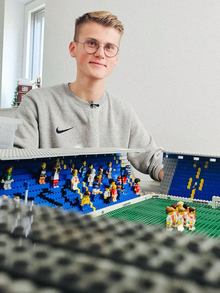 Ein junger Mann in grauem Pulli und kurzen blonden Haaren sitzt hinter einem aus Lego gebauten Fußballstadion