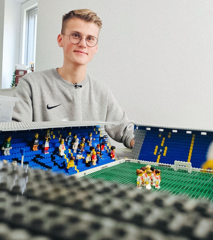 Ein junger Mann in grauem Pulli und kurzen blonden Haaren sitzt hinter einem aus Lego gebauten Fußballstadion