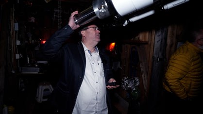 Hubertus Rieger schaut durch ein großes Teleskop. Mit seiner rechten Hand stützt er sich dafür an dem weiß-schwarzen Teleskop ab.Er trägt ein weißes Hemd und eine schwarze Jacke. 