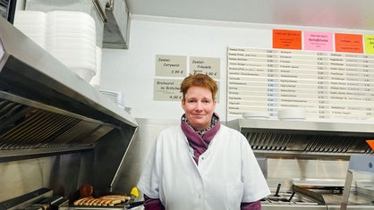 Eine Frau mit kurzen roten Haaren und weißem Kittel steht hinter einer Theke und lächelt