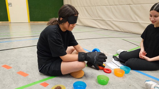 Ein Mädchen sitzt auf dem Boden einer Sporthalle, trängt Winterhandschuhe und eine Augenmaske und ertastet Gegenstände.