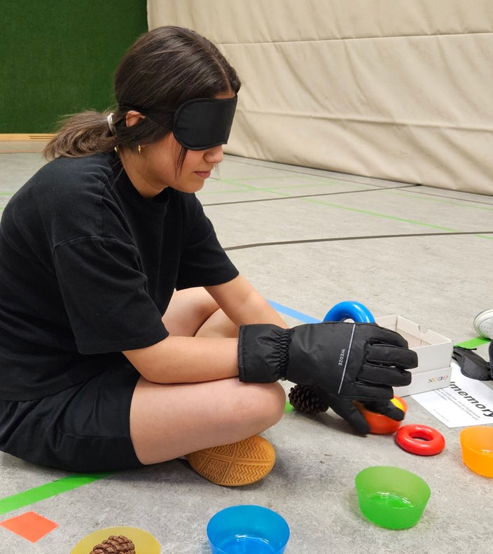 Ein Mädchen sitzt auf dem Boden einer Sporthalle, trängt Winterhandschuhe und eine Augenmaske und ertastet Gegenstände.