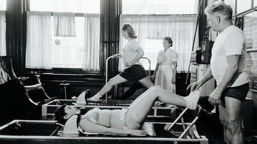 Joseph Pilates unterstützt eine Sportlerin beim Training an einem Gerät.