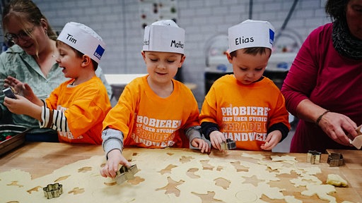 Drei Kinder mit Papierhütchen stanzen mit Formen Keksteig.