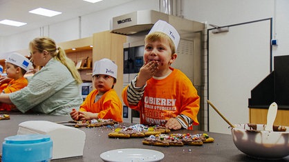 Mehrere Kinder dekorieren Keks-Weihnachtsbäume, dabei nascht einer der Kinder und hat ein mit Schokolade verschmiertes Gesicht.