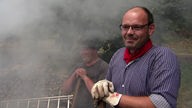 Hobbyköhler Christof Kordes lächelt, im Hintergrund ziehen Rauchschwaden vom Kohlemeiler.