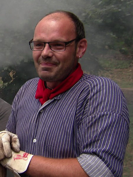 Hobbyköhler Christof Kordes lächelt, im Hintergrund ziehen Rauchschwaden vom Kohlemeiler.