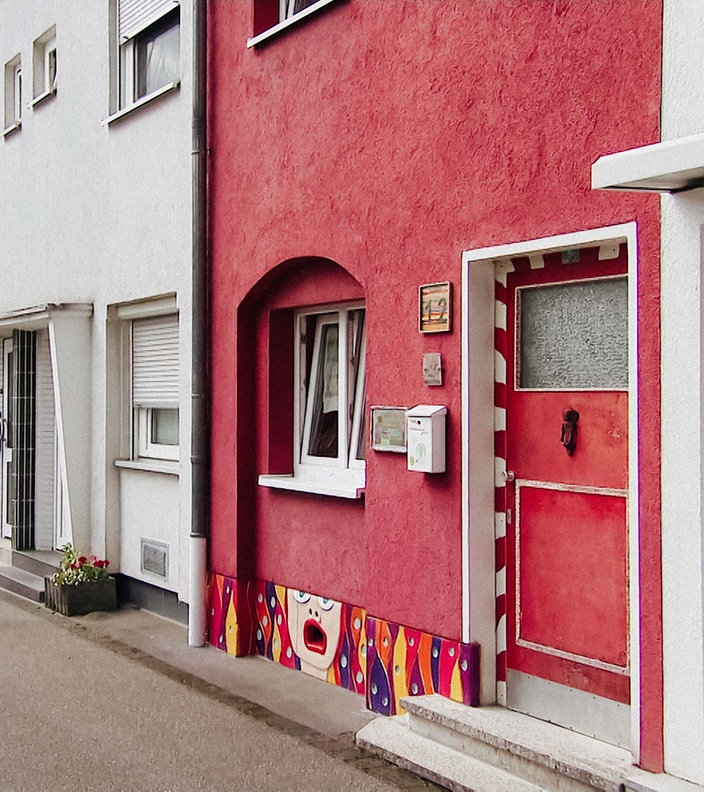 Ein Haus in einer Straße ist rot gestrichen, am Sockel ist ein buntes Motiv gemalt.