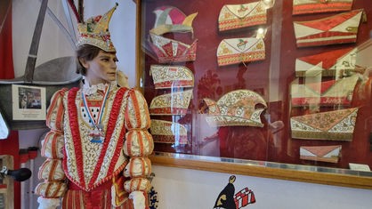 Eine Schaufensterpuppe, die das Kostüm eines Karnevalsprinzen trägt, daneben eine hängende Vitrine mit Karnevalsmützen im Karnevalsmuseum in Köln-Dünnwald.