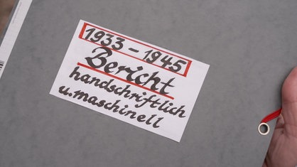 Ein grauer Pappordner, auf dem in säuberlicher, alter Handschrift: "1933-1945 Bericht handschriftlich u. maschinell" steht