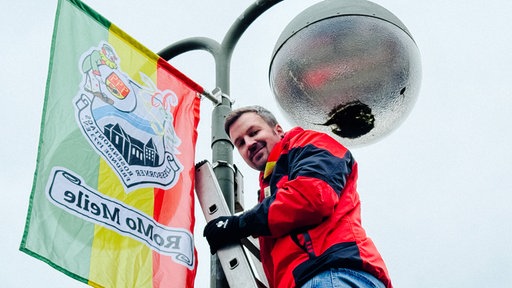 Markus Bismor ist mit einer Leiter auf eine Laterne geklettert und schmückt diese nun mit einer Karnevalsflagge