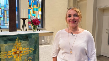 Eine blonde Frau, die vor einem Altar steht und in die Kamera lächelt